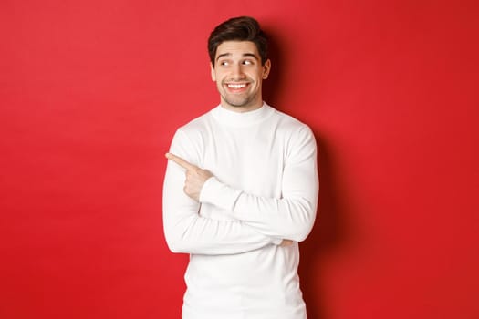 Concept of winter holidays, christmas and lifestyle. Excited handsome man in white sweater, smiling, looking and pointing finger left at promo offer, standing over red background.