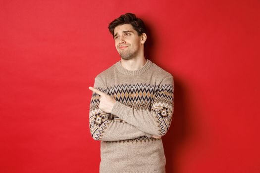 Image of disappointed handsome man in christmas sweater, looking and pointing left with upset face, express regret, showing new year advertisement, standing over red background.