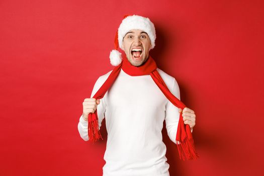 Image of angry adult man hate christmas, wearing scarf and santa hat, yelling distressed, standing mad over red background.