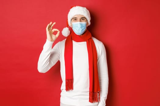 Concept of covid-19, christmas and holidays during pandemic. Portrait of happy and satisfied man in medical mask and santa hat, showing okay sign in approval, recommending something.