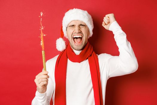 Concept of christmas, winter holidays and celebration. Handsome man celebrating new year and having fun, holding sparkler and smiling, wearing santa hat, standing over red background.