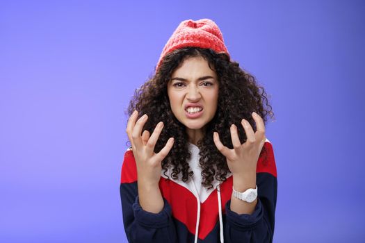 Pissed and annoyed stylish girl clenching hands in anger, grimacing squeezing teeth from annoyance and irritation, standing hateful and outraged feeling fury and hate posing over blue background.