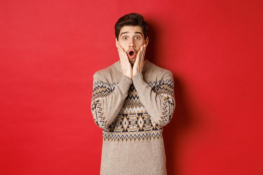 Image of surprised handsome guy reacting to cool new year promo offer, gasping amazed, wearing christmas sweater, standing over red background.