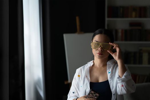 Asian middle aged woman is disco shades or party glasses with easel behind her. Mature serious looking at camera woman, Happy feeling concept.