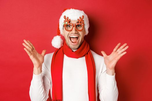 Concept of christmas, winter holidays and celebration. Image of surprised and happy man looking amazed, wearing party glasses and enjoying new year, standing over red background.