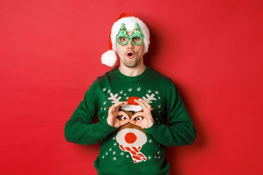 Portrait of carefree handsome man in santa hat and party glasses, making fun of his christmas sweater, looking happy over red background.