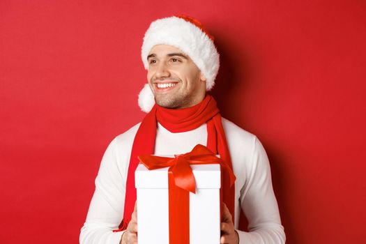Concept of winter holidays, christmas and lifestyle. Close-up of attractive guy in santa hat and scarf, smiling and looking left while holding new year present, standing over red background.