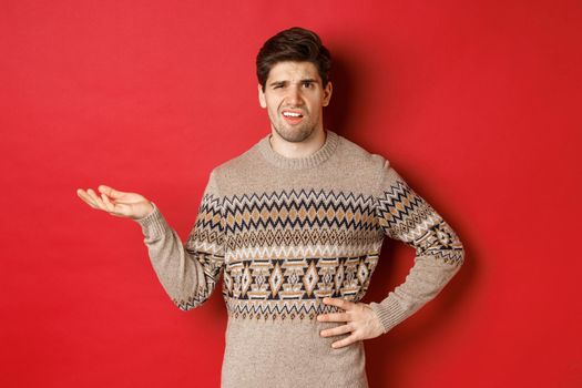 Image of confused and disappointed handsome man, complaining about christmas party, raising hand and looking bothered, standing over red background.