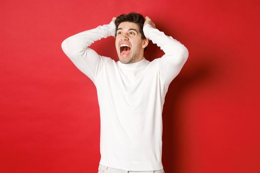 Portrait of frustrated man in white sweater, screaming in panic and looking at upper left corner, have terrible problem, standing over red background.