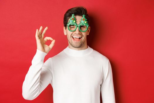 Portrait of handsome and cheeky guy, wearing party glasses and white sweater, showing okay sign and winking, wishing happy new year, standing over red background.