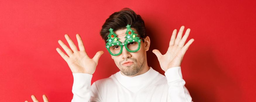Concept of winter holidays, christmas and celebration. Close-up of funny handsome guy in party glasses, showing empty hands, standing over red background.