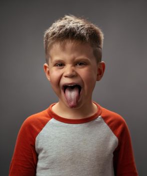 Sticking out tongue boy disobedience, bad behavior. Funny laughing boy in red shirt with eyes shut. Happy kid on dark grey background. Funny family time. Little boy enjoying having fun in studio.