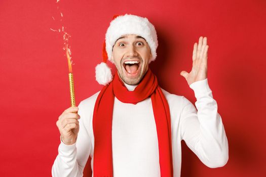 Concept of christmas, winter holidays and celebration. Handsome man celebrating new year and having fun, holding sparkler and smiling, wearing santa hat, standing over red background.