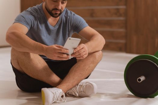 Body shot of handsome young man searching online or texting sitting on floor with smartphone, black and green tone fitness barbell, equipment for weight training concept. Healthy lifestyle concept.