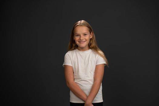 Smiling girl looking timid or frustrated at the camera wearing white t-shirt isolated on dark grey or black background. Child emotions concept.