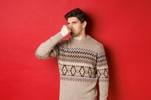 Image of disgusted young man in winter sweater, shut nose and grimacing from bad smell, standing over red background.