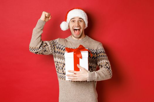 Image of happy and excited handsome man, reicing christmas gift, raising hands up in triumph and smiling, celebrating new year, standing over red background in santa hat.
