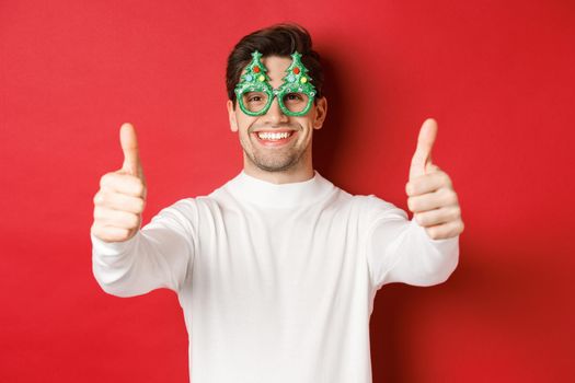 Close-up of attractive happy man in party glasses and white sweater, showing thumbs-up in approval and smiling, standing over red background.