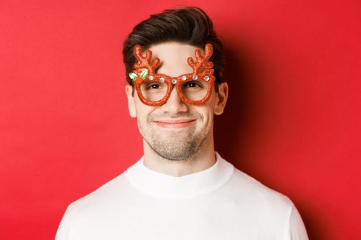 Concept of winter holidays, christmas and celebration. Close-up of attractive smiling male model in party glasses, standing against red background.