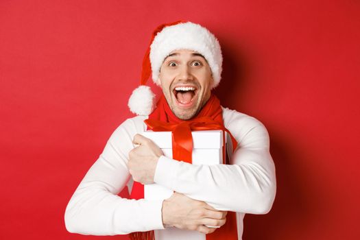 Concept of winter holidays, christmas and lifestyle. Close-up of happy man in santa hat receiving present, looking happy and hugging gift box, standing over red background.
