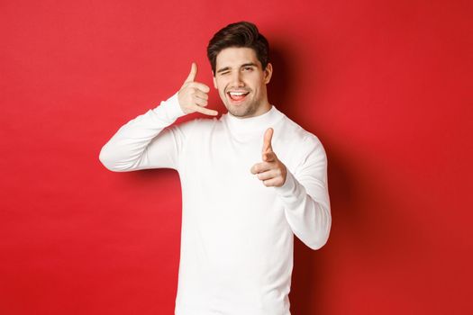 Portrait of sassy and confident handsome man, flirting with you, asking to call him, showing phone sign and pointing at camera, standing over red background.