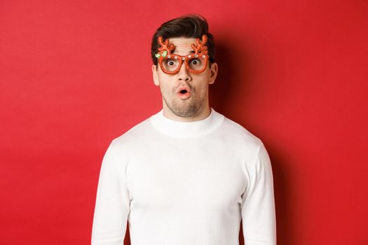 Close-up of surprised and impressed man in white sweater, party glasses, saying wow and looking amazed at camera, standing over red background.
