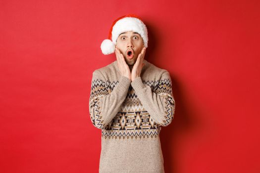 Image of amazed and speechless man in santa hat, drop jaw and staring at awesome christmas offer, standing over red background.