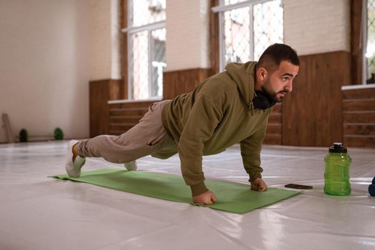 Muscular sportsman doing exercises alone in gym. Young handsome sports guy doing push ups exercise in empty gym with green bottle of water next to him all in khaki color clothes.