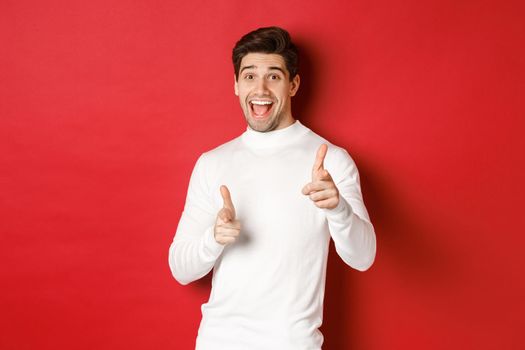 Concept of winter holidays, christmas and lifestyle. Cheerful handsome guy in white sweater congratulating you, pointing fingers at camera and wishing happy new year, standing over red background.