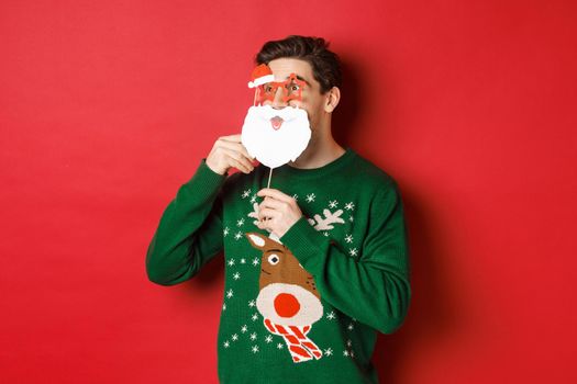 Portrait of surprised man in green christmas sweater, holding funny santa claus mask, celebrating new year, looking left amazed, sanding over red background.