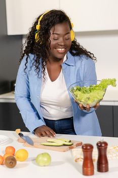 Afro Female Listens to Music With Headphones and Makes a Salad For a Breakfast in the Kitchen at Home. A Fresh Salad in Glass Bowl . Close- up. High quality photo