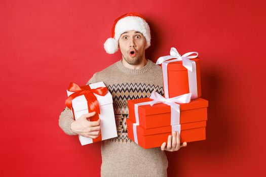 Concept of winter holidays, new year and celebration. Image of surprised handsome guy in sweater and santa hat, receiving gifts, holding boxes with presents and looking amazed.