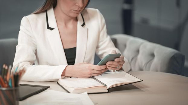 Close up beautiful office worker in white jacket making notes in her journal holding smartphone or phone in her hands sitting in front of journal. Office worker looking at her working place.