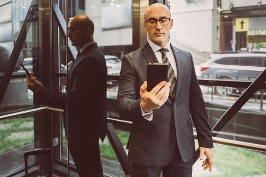 Thoughtful businessman in suit reading information in smartphone hold in it in front of him standing next to a window with a busy street view. Focused business man face with smartphone.