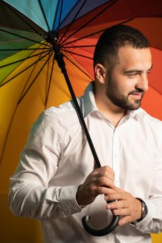 A nice guy with a stylish beard and white shirt stands with a rainbow umbrella behind his back. Handsome guy in support of LGBT society