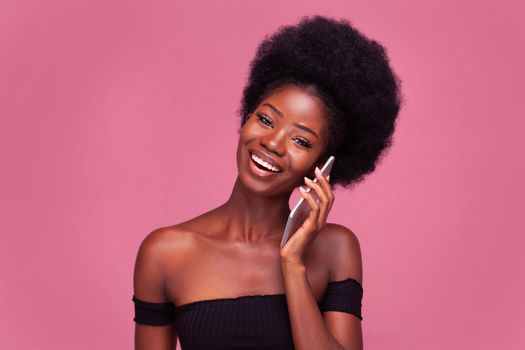 Cheerful talking on the phone African American woman chatting with girlfriends using her smartphone. Female model with afro hairstyle in black cut top with bared shoulders isolated on pink.