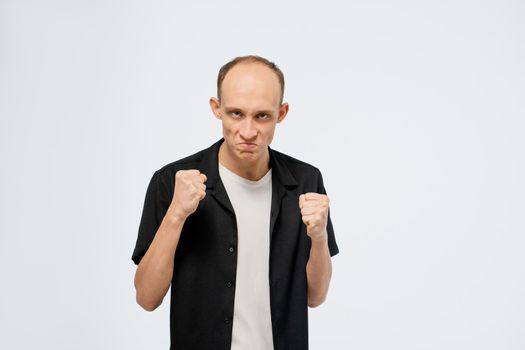 Defending himself stands in a stance with ready fists young bald man in black shirt with white t-shirt under. Angry young adult man showing he is ready for fight isolated on white background.