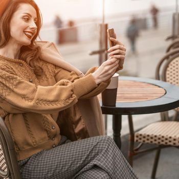 Beautiful young woman video calling using her smartphone while having cup of coffee sitting in a spring outdoor cafe. French woman in red beret with background of urban city. Square cropped.