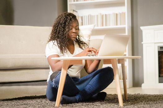 Pretty Afro Girl With Folded Legs Sitting on the Carpet and Typing on her Computer. African Girl Looks Through the List of Films on Her Laptop. Close-up. Home Background . High quality photo