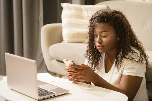 African Woman in White T-shirt is Sitting on the Floor and Texting a Message on Her Smartphone Next to Her Laptop. Close-up. Couch Background. High quality photo