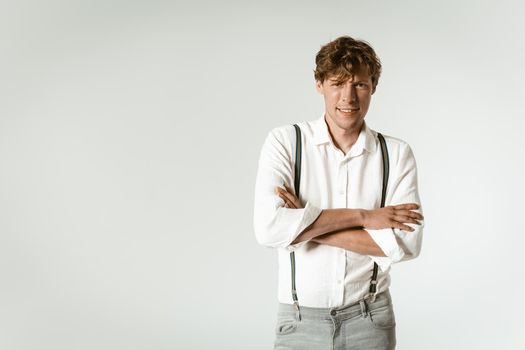 Handsome young hipster man with his arms crossed posing with hands folded dressed in white shirt and grey jeans isolated on white background. Copy space at left side.