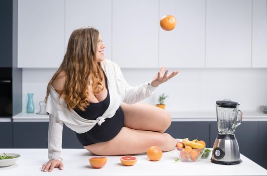 An obese young sexy chubby white girl in black swimsuit and white shirt at modern kitchen with fruits next to her. Sexy overweight girl laying on the kitchen table.