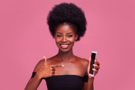 Call me. Cheerful African American young girl pointing finger at phone saying call me with gorgeous afro hairstyle standing smiling in black shoulder cut top isolated on pink background.