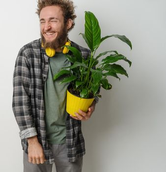 Handsome smiling young bearded man holding yellow flower pot with plant dressed in plaid shirt and yellow headphones on his neck isolated on white background. Moving concept.