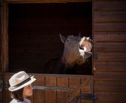 Charming young girl in vintage clothes standing with her brown horse sticking out head of the barn the horse happy seeing its human friend. Friendship concept.