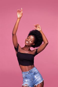 Dancing with hands up gorgeous young African American girl with afro hair posing dressed in black top and denim jeans on dirty pink background.