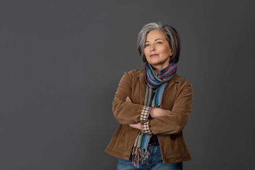 Pretty grey hair woman in casual isolated on grey background. Portrait of mature grey haired woman in a brown jacket looking positive on camera.