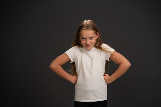 Bossy girl with her hands sideways look at the camera wearing white t shirt isolated on dark grey or black background.