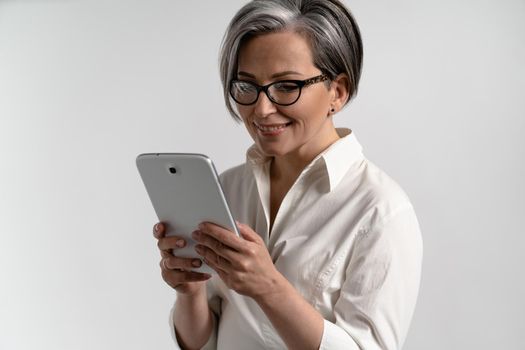 Portrait of a grey haired senior woman in white shirt and eye glasses using digital tablet. Technology by the elderly browsing internet shopping online using pad isolated on white background.