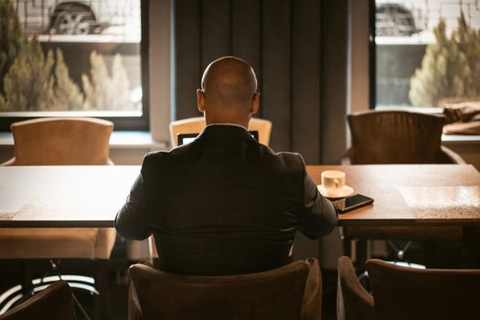 Back view of elegant CEO dressed in expensive suit sitting at the table in the modern expensive office interior, bald male working on laptop. Back view.